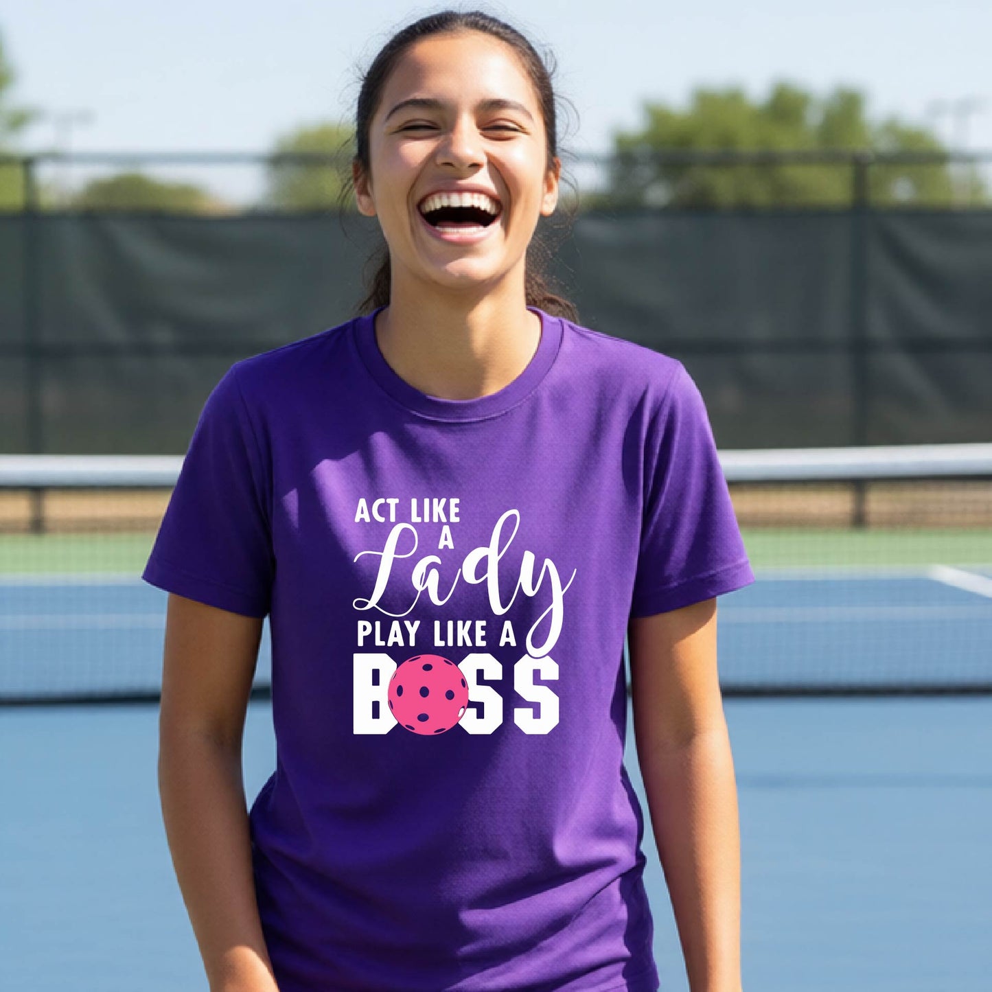 A young person laughing on an outdoor pickleball court wearing a purple t-shirt with a graphic that reads 'ACT LIKE A Lady PLAY LIKE A BOSS' with a pink pickleball graphic.