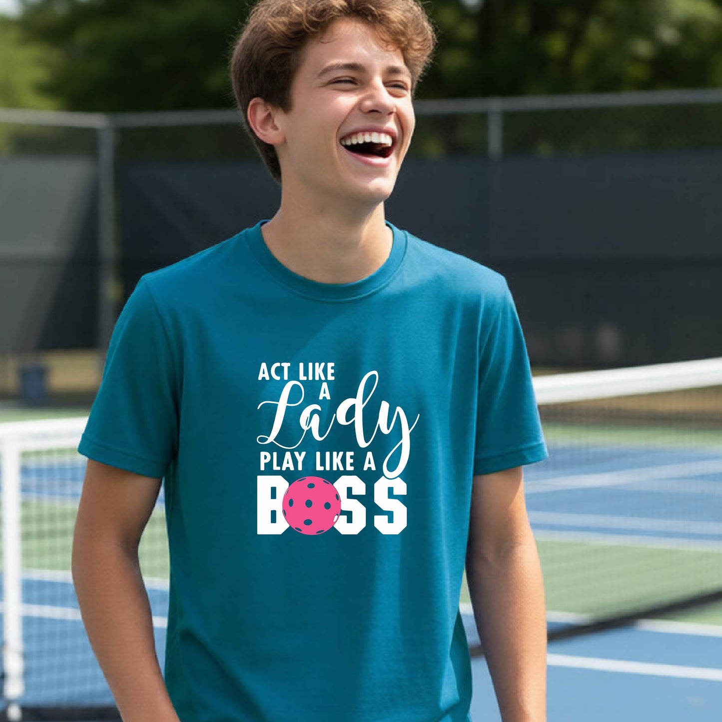 A young person laughing on an outdoor pickleball court wearing a teal t-shirt with a graphic that reads 'ACT LIKE A Lady PLAY LIKE A BOSS' with a pink pickleball graphic.
