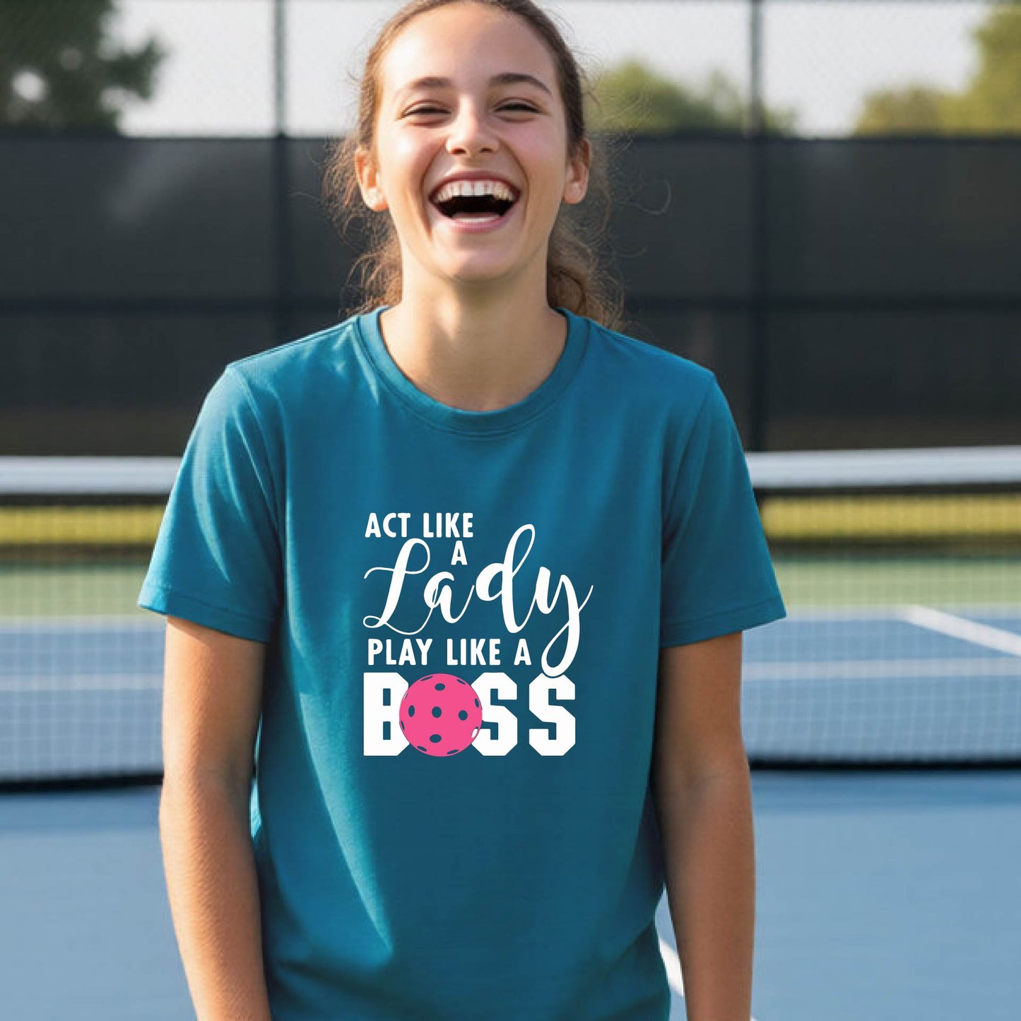 A young person laughing on an outdoor pickleball court wearing a teal t-shirt with a graphic that reads 'ACT LIKE A Lady PLAY LIKE A BOSS' with a pink pickleball graphic.