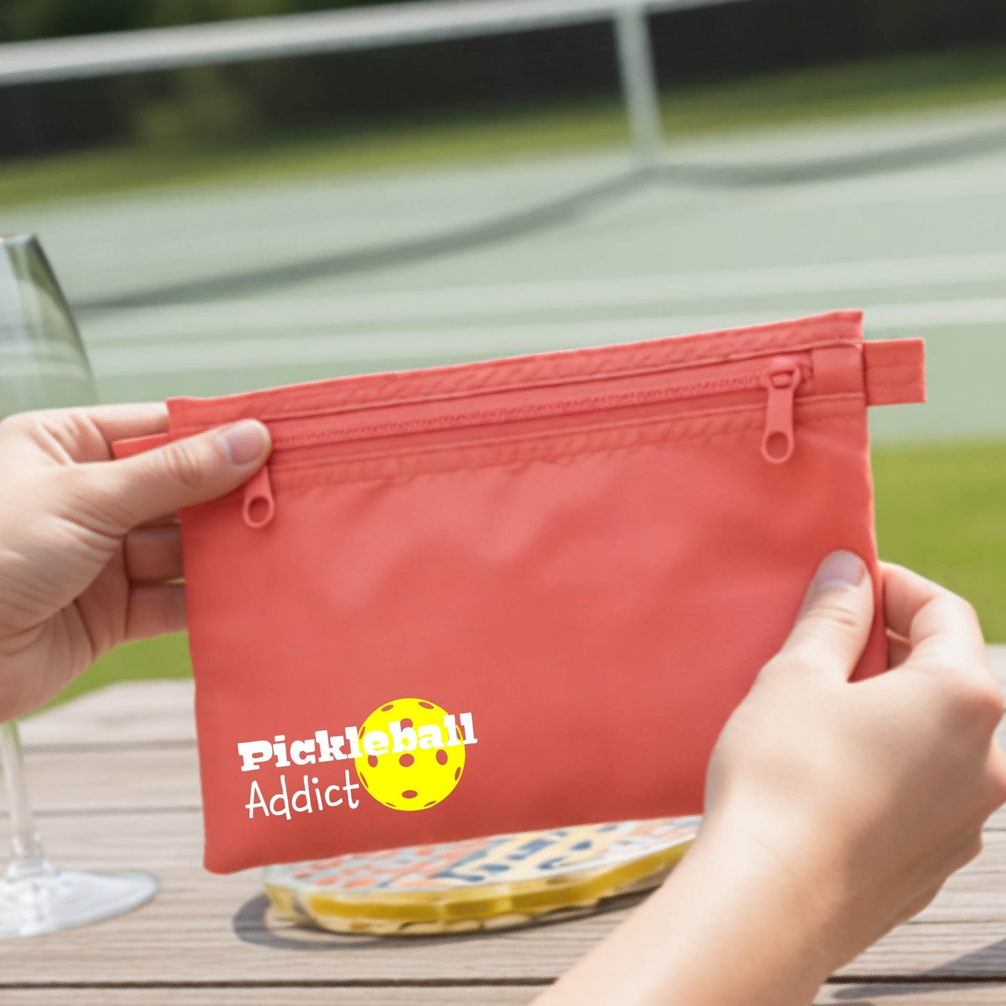 A coral red zippered pouch with 'Pickleball Addict' printed on the front, with a blurred pickleball court in the background.