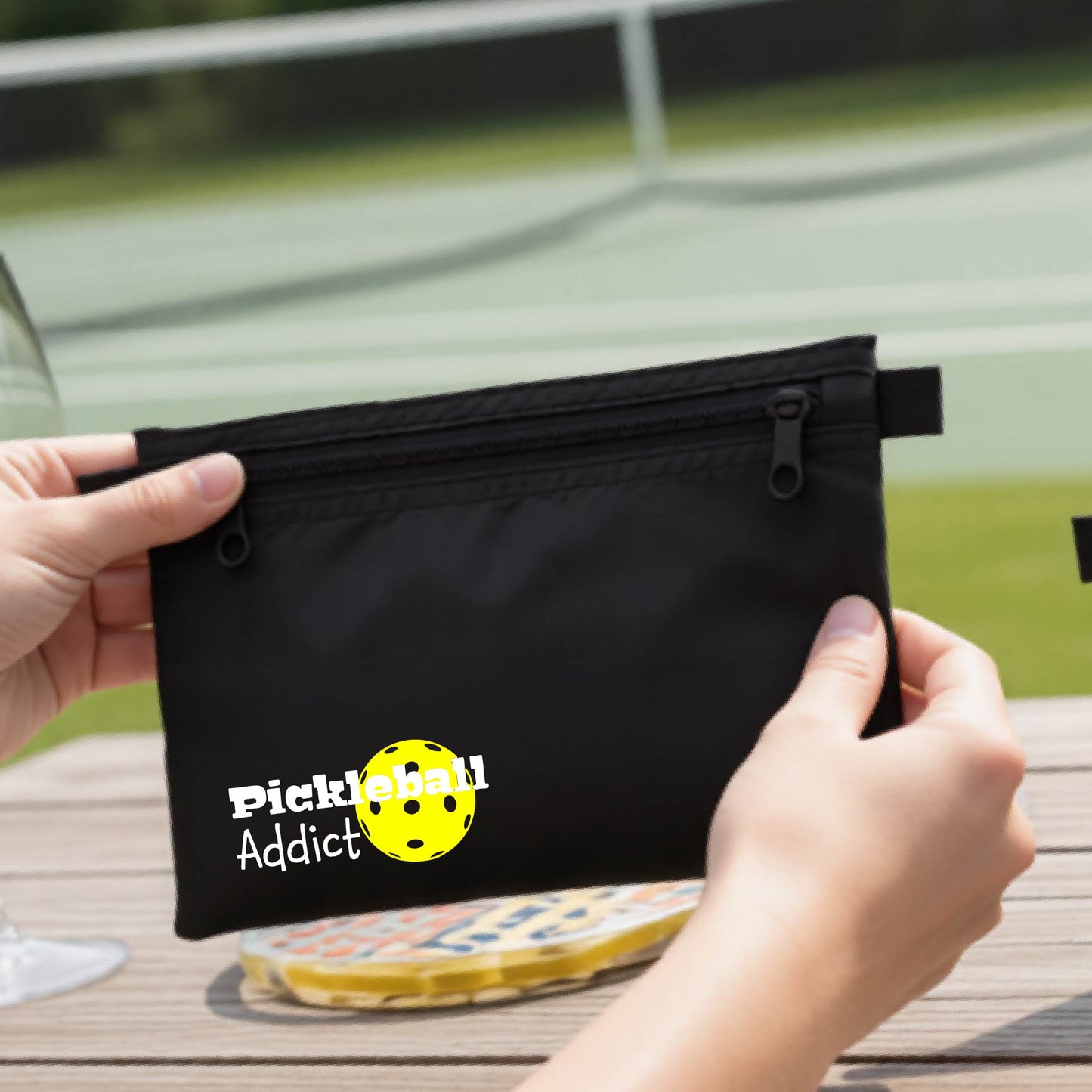 A black zippered pouch with 'Pickleball Addict' printed on the front, with a blurred pickleball court in the background.