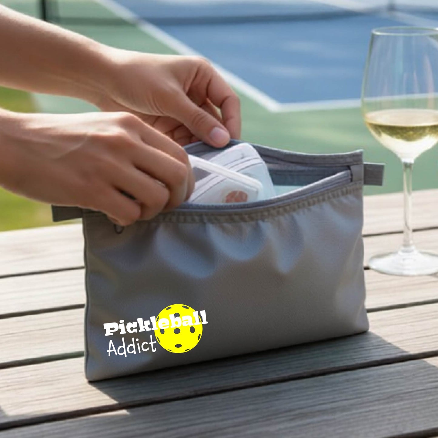 A gray zippered pouch with 'Pickleball Addict' printed on the front, with a blurred pickleball court in the background.