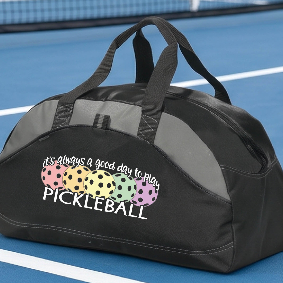 A black and gray pickleball duffel bag with gray panels, on a green pickleball court . The bag features a graphic of colorful pickleballs and the text 'it's always a good day to play PICKLEBALL'.