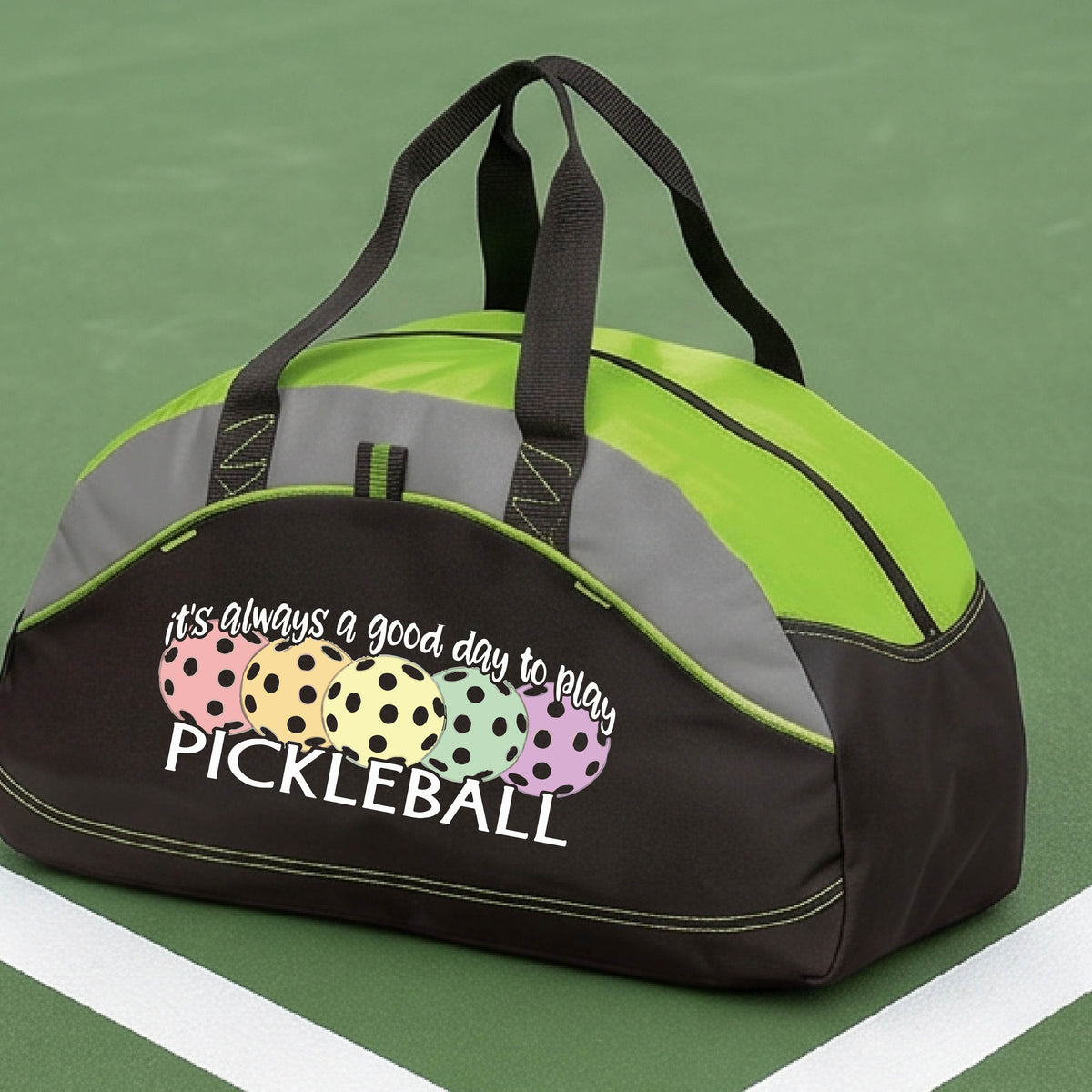 A black and green lime pickleball duffel bag with gray panels, on a green pickleball court . The bag features a graphic of colorful pickleballs and the text 'it's always a good day to play PICKLEBALL'.