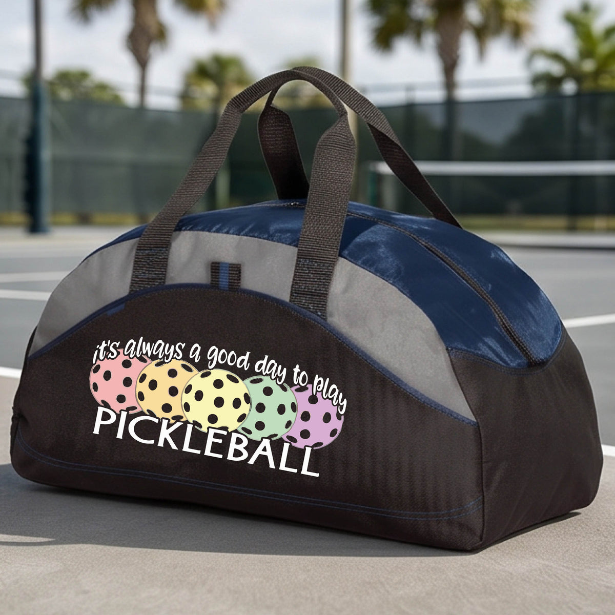 A black and navy blue pickleball duffel bag with gray panels, on a green pickleball court . The bag features a graphic of colorful pickleballs and the text 'it's always a good day to play PICKLEBALL'.