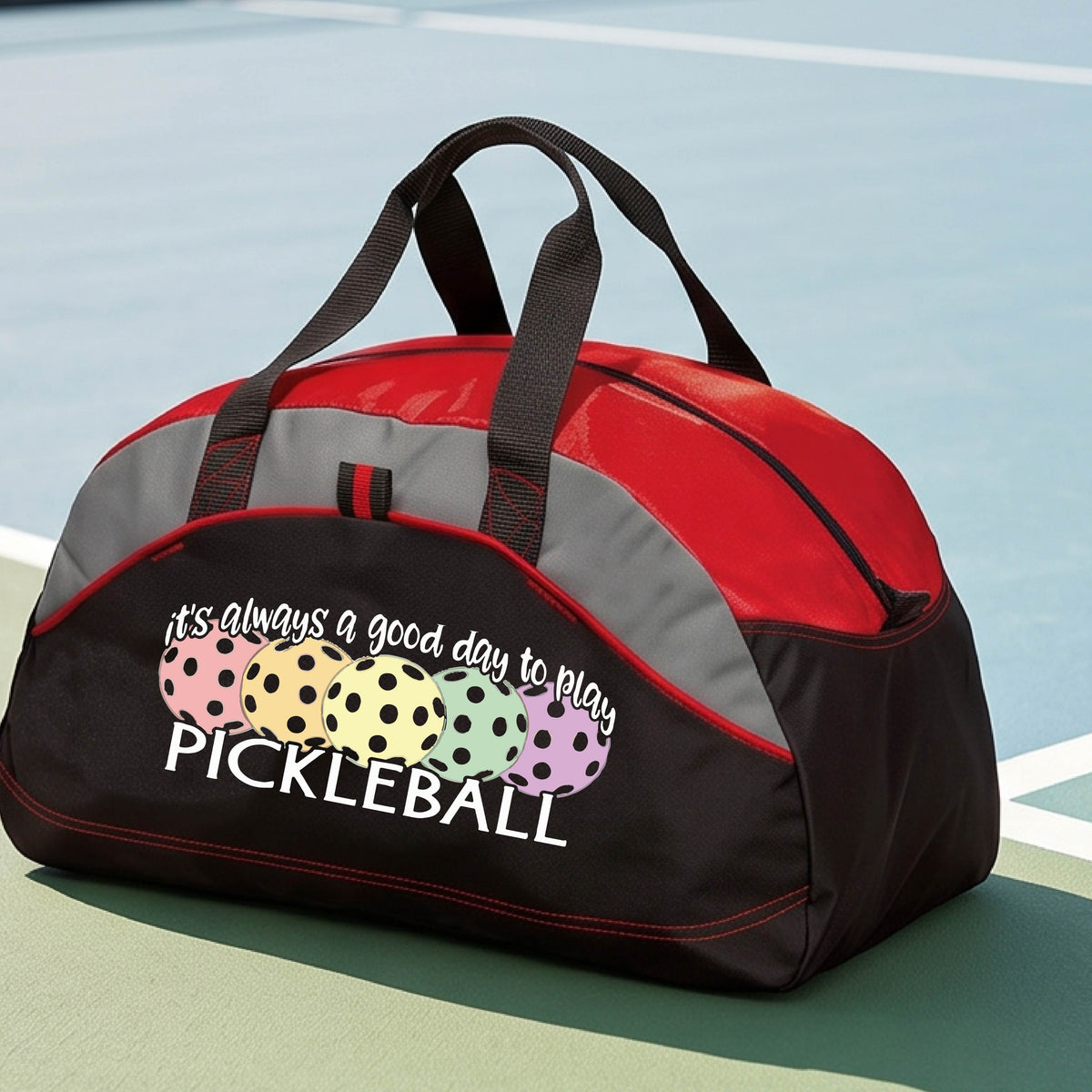 A red, black and gray pickleball duffel bag with gray panels, on a green pickleball court . The bag features a graphic of colorful pickleballs and the text 'it's always a good day to play PICKLEBALL'.