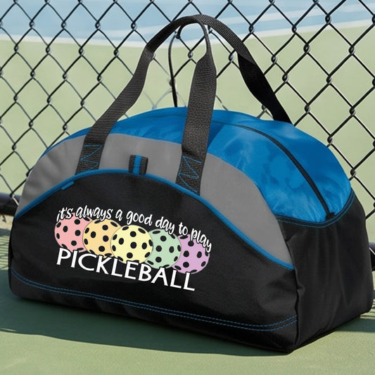A black and blue pickleball duffel bag with gray panels, on a green pickleball court . The bag features a graphic of colorful pickleballs and the text 'it's always a good day to play PICKLEBALL'.