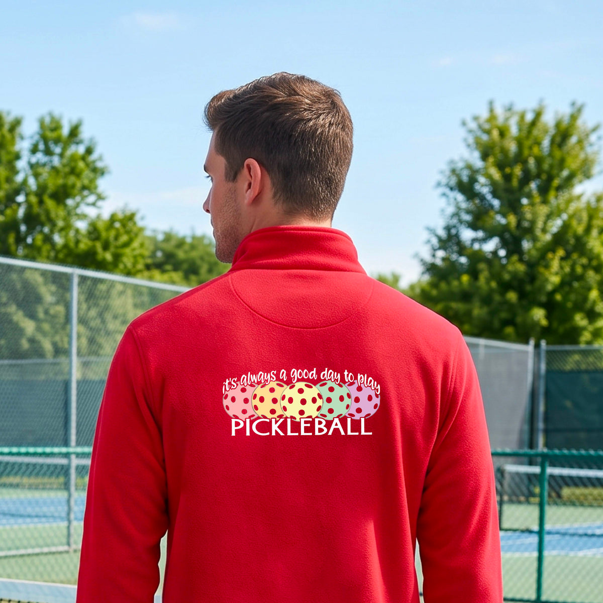 A man on a pickleball court seen from behind, wearing a red long-sleeve quarter-zip athletic shirt with a colorful graphic on the back that reads 'it's always a good day to play PICKLEBALL' above and below three  pickleball balls.