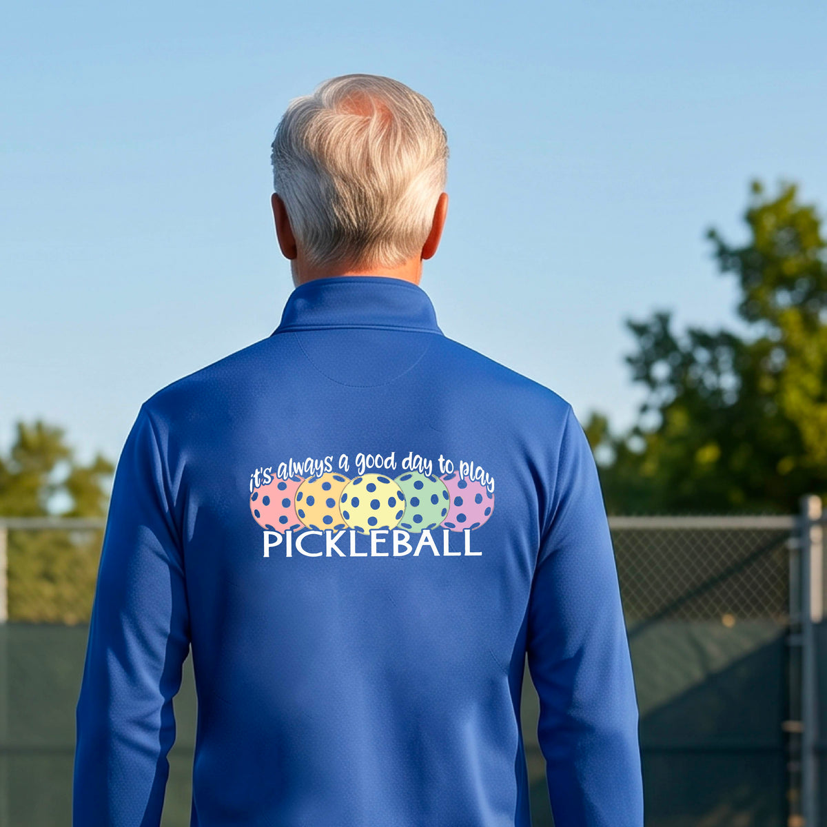 A man on a pickleball court seen from behind, wearing a royal blue long-sleeve quarter-zip athletic shirt with a colorful graphic on the back that reads 'it's always a good day to play PICKLEBALL' above and below three  pickleball balls.