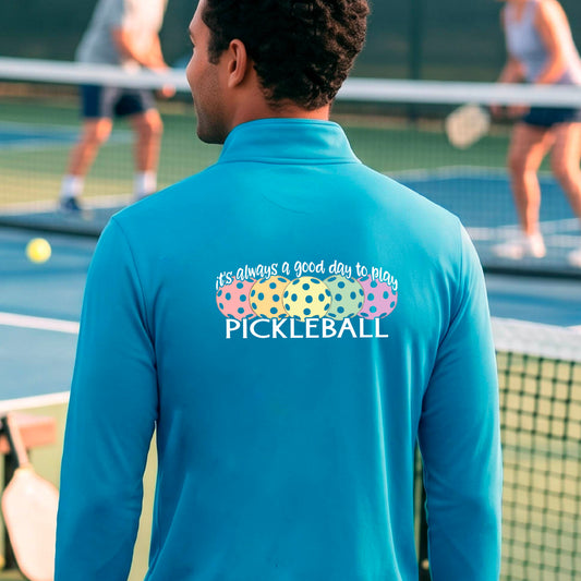 A man on a pickleball court seen from behind, wearing a bright turquoise long-sleeve quarter-zip athletic shirt with a colorful graphic on the back that reads 'it's always a good day to play PICKLEBALL' above and below three  pickleball balls.