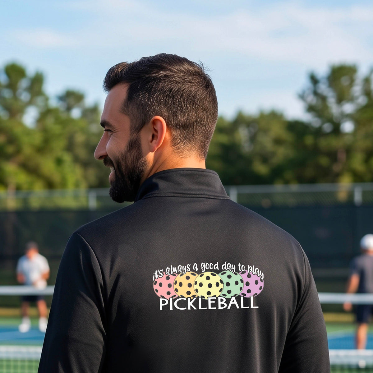 A man on a pickleball court seen from behind, wearing a black long-sleeve quarter-zip athletic shirt with a colorful graphic on the back that reads 'it's always a good day to play PICKLEBALL' above and below three  pickleball balls.
