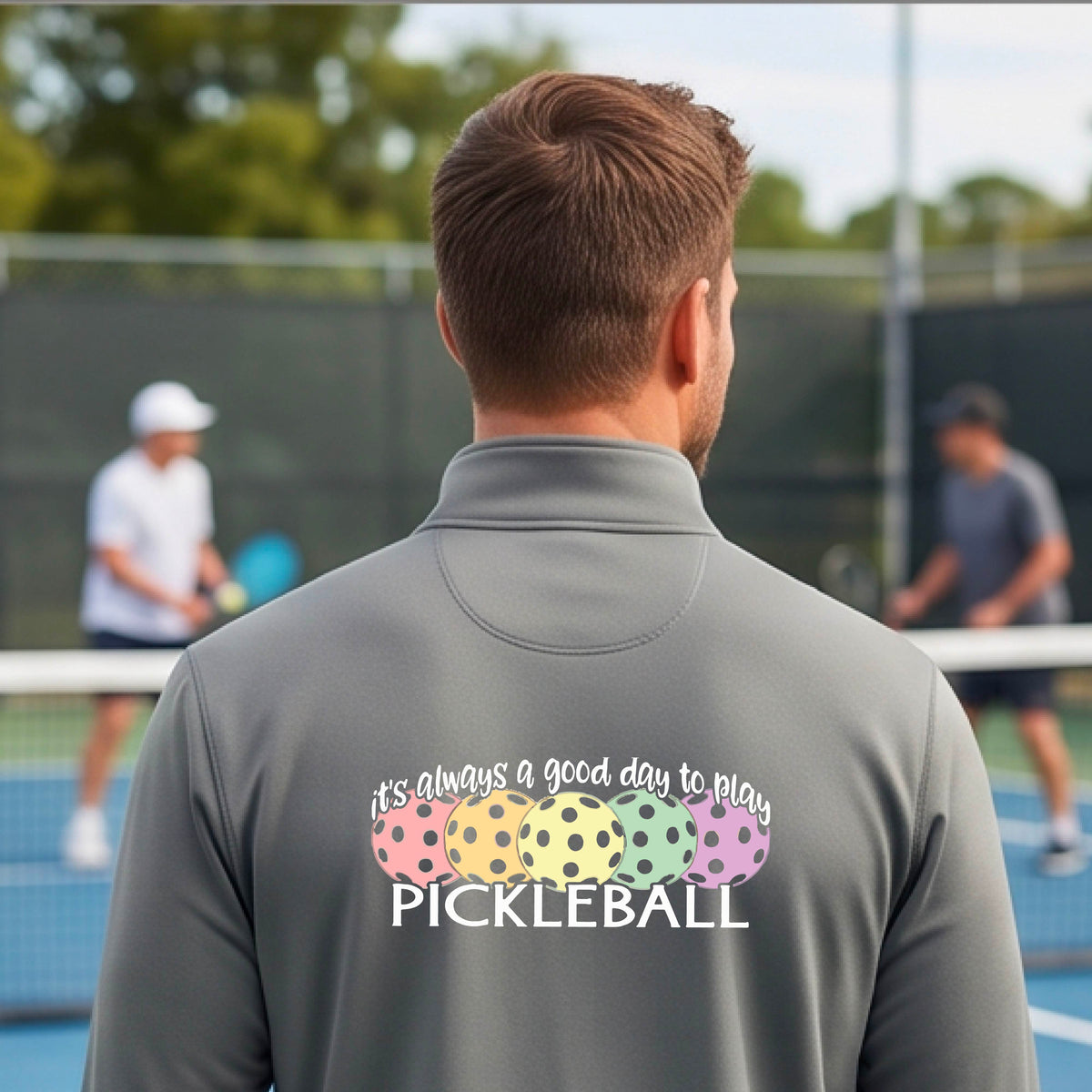 A man on a pickleball court seen from behind, wearing a gray long-sleeve quarter-zip athletic shirt with a colorful graphic on the back that reads 'it's always a good day to play PICKLEBALL' above and below three  pickleball balls.