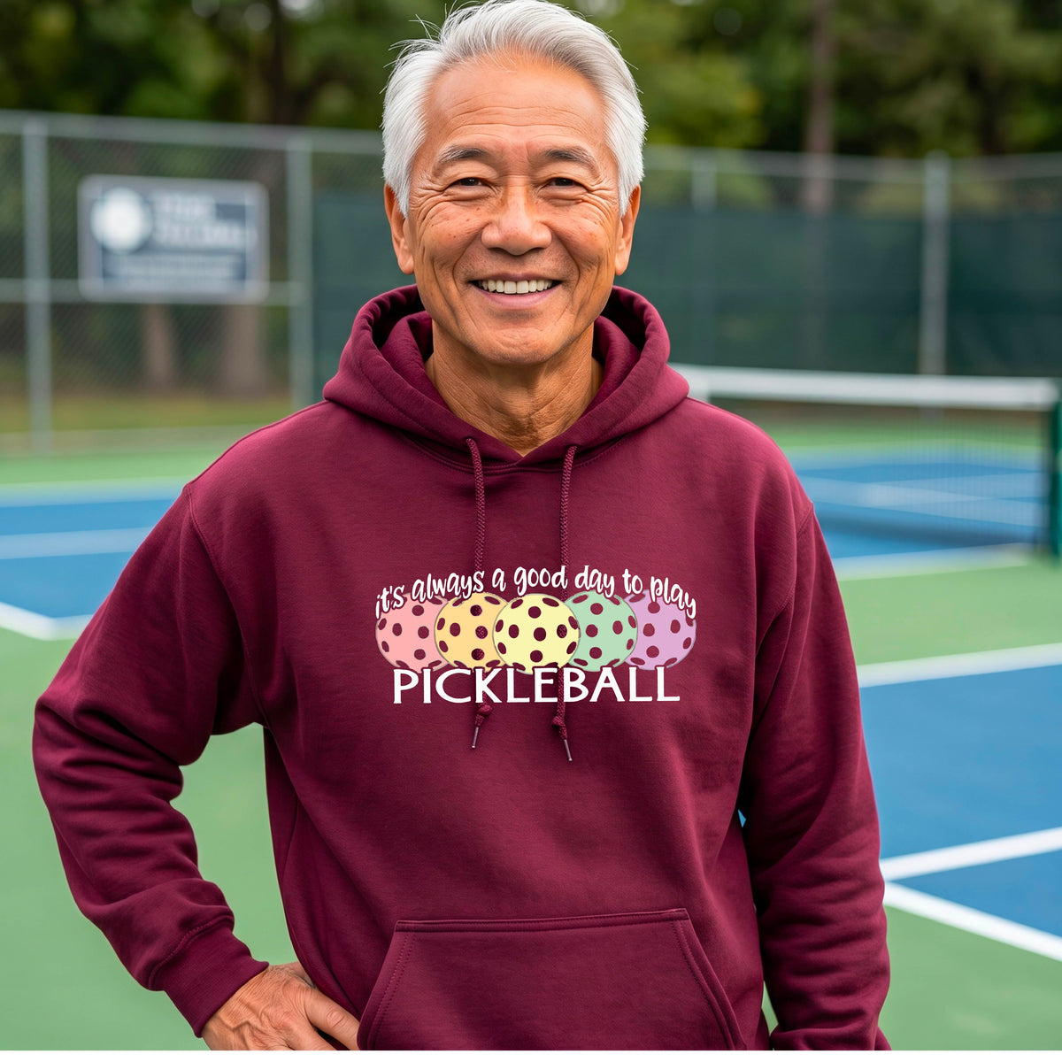 A man smiling outdoors wearing a maroon hoodie with a colorful pickleball graphic and the text 'it's always a good day to play PICKLEBALL' on the chest.
