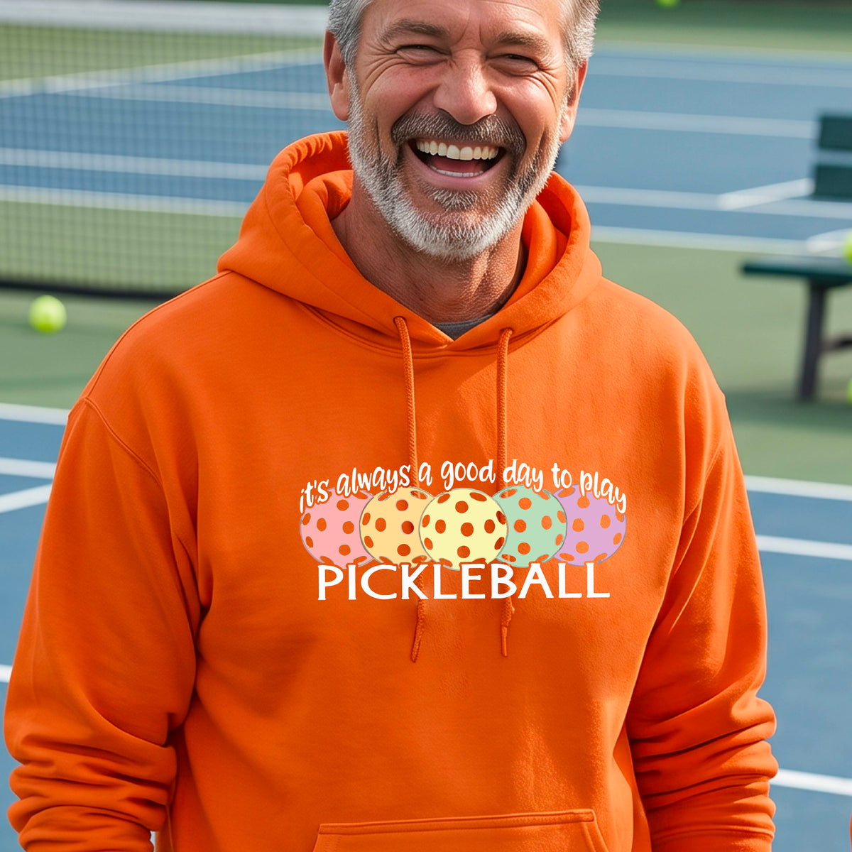 A man smiling outdoors wearing an orange hoodie with a colorful pickleball graphic and the text 'it's always a good day to play PICKLEBALL' on the chest.