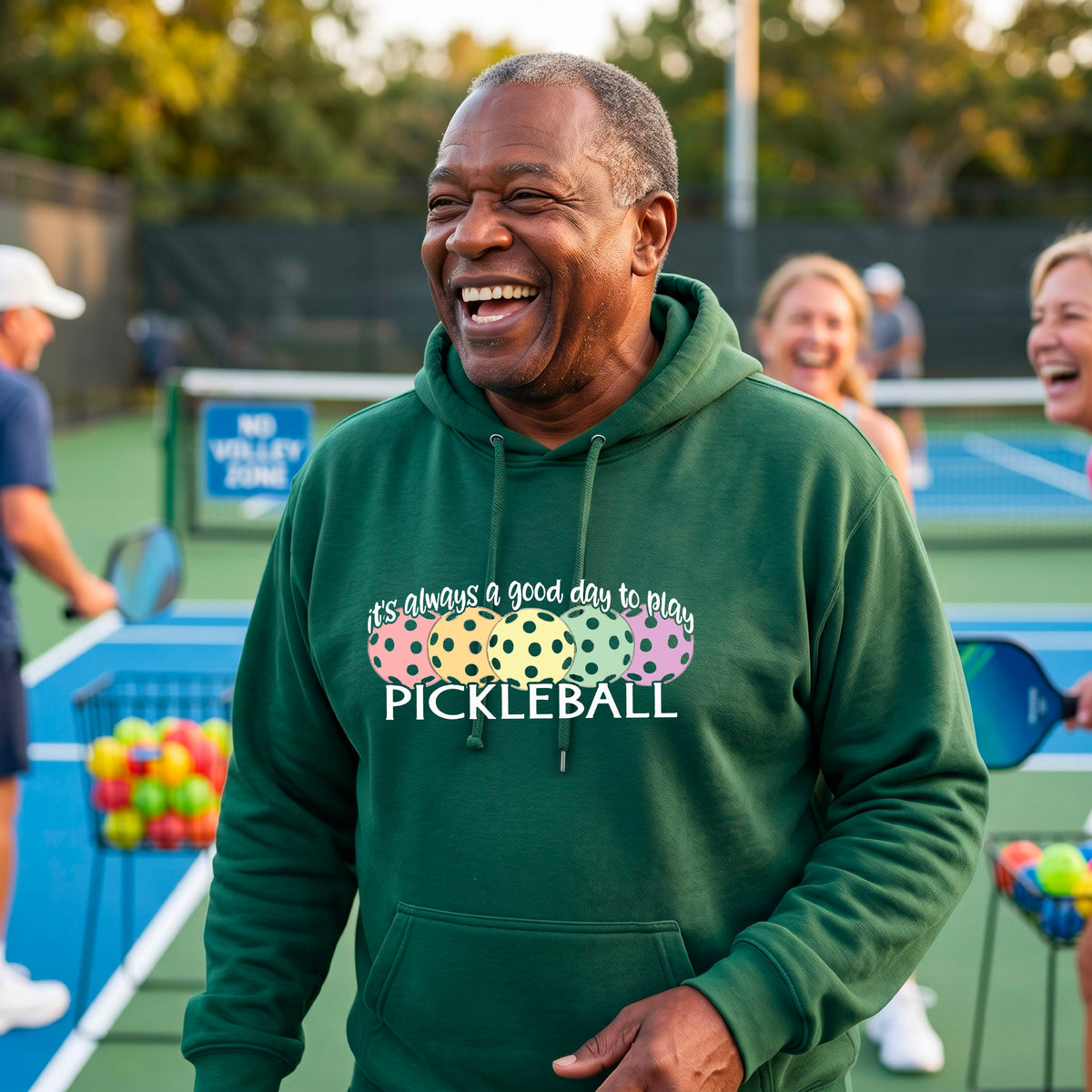 A man smiling outdoors wearing a green hoodie with a colorful pickleball graphic and the text 'it's always a good day to play PICKLEBALL' on the chest.