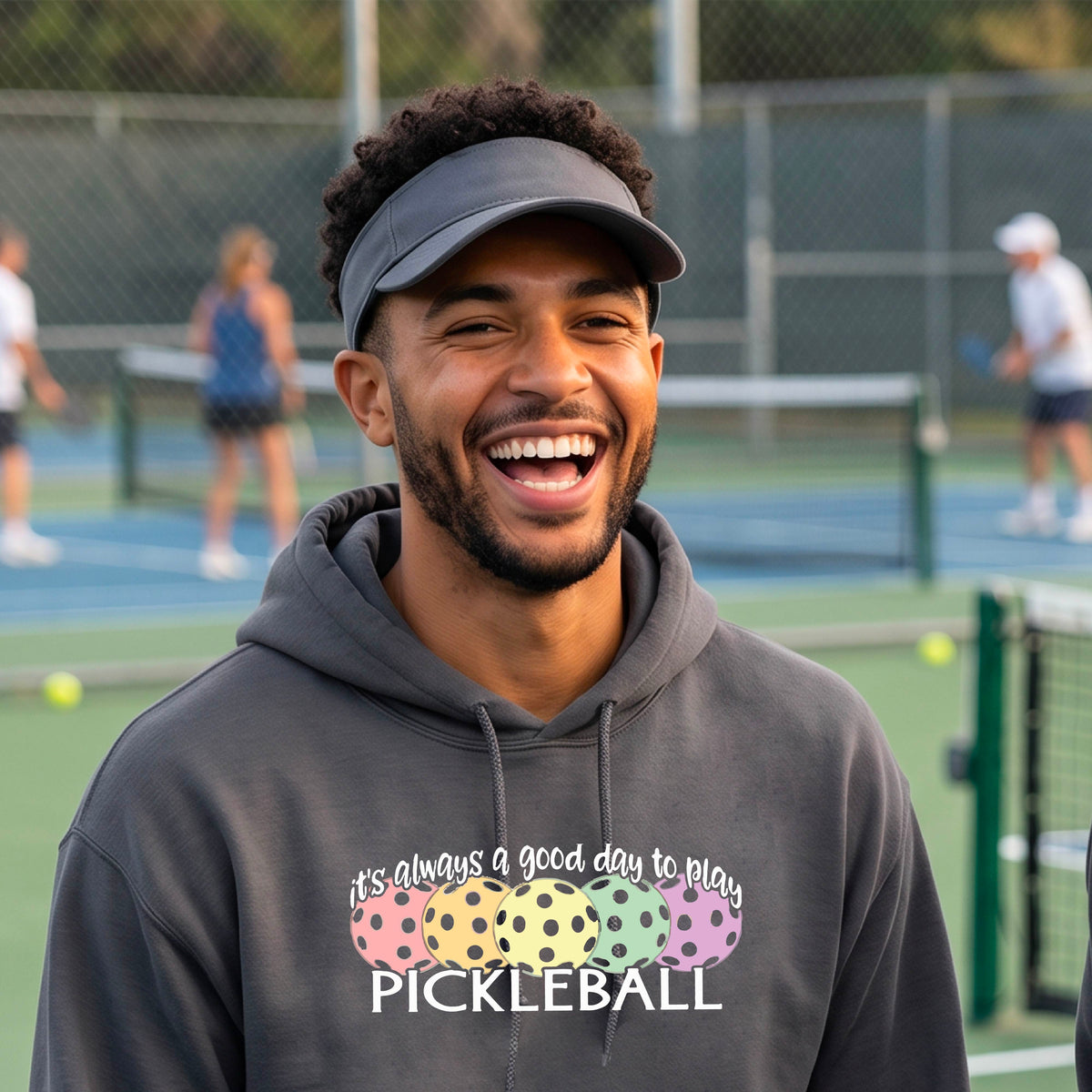 A man smiling outdoors wearing a gray hoodie with a colorful pickleball graphic and the text 'it's always a good day to play PICKLEBALL' on the chest.