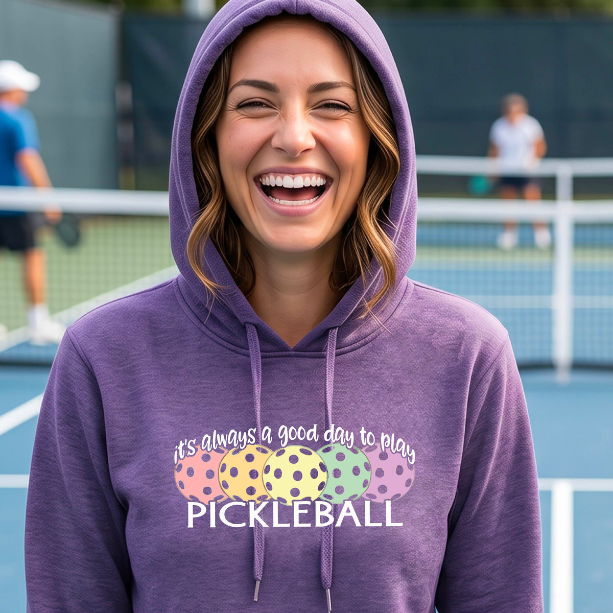 A woman smiling on a pickleball court wearing a purple hoodie with a graphic that reads 'it's always a good day to play PICKLEBALL' above four colorful pickleballs.
