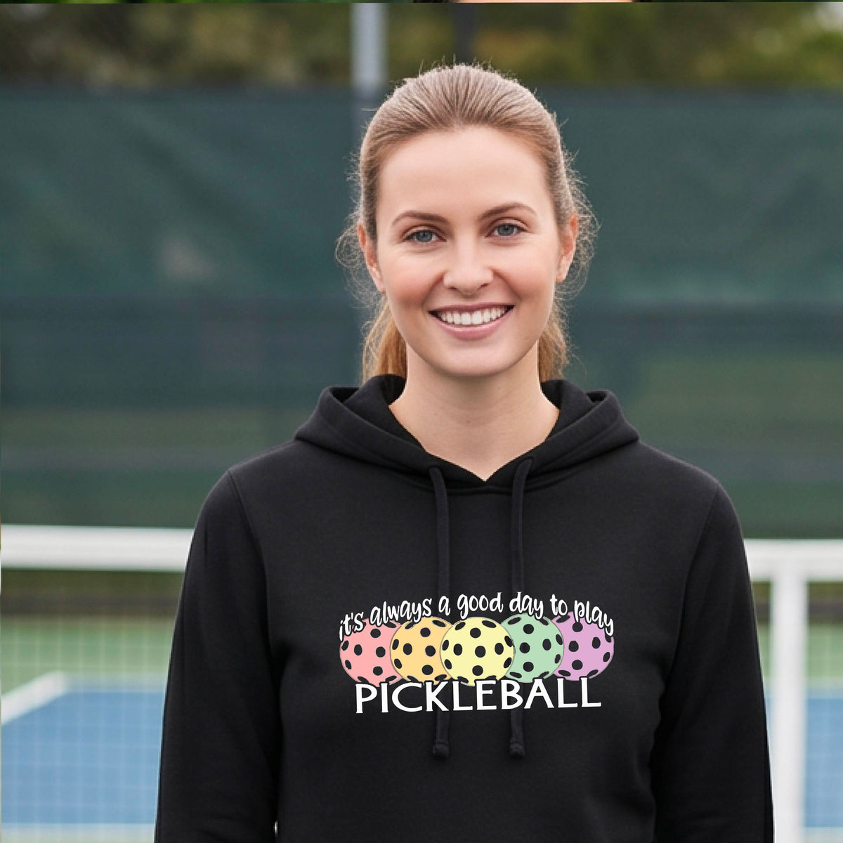 A woman smiling on a pickleball court wearing a black hoodie with a graphic that reads 'it's always a good day to play PICKLEBALL' above four colorful pickleballs.