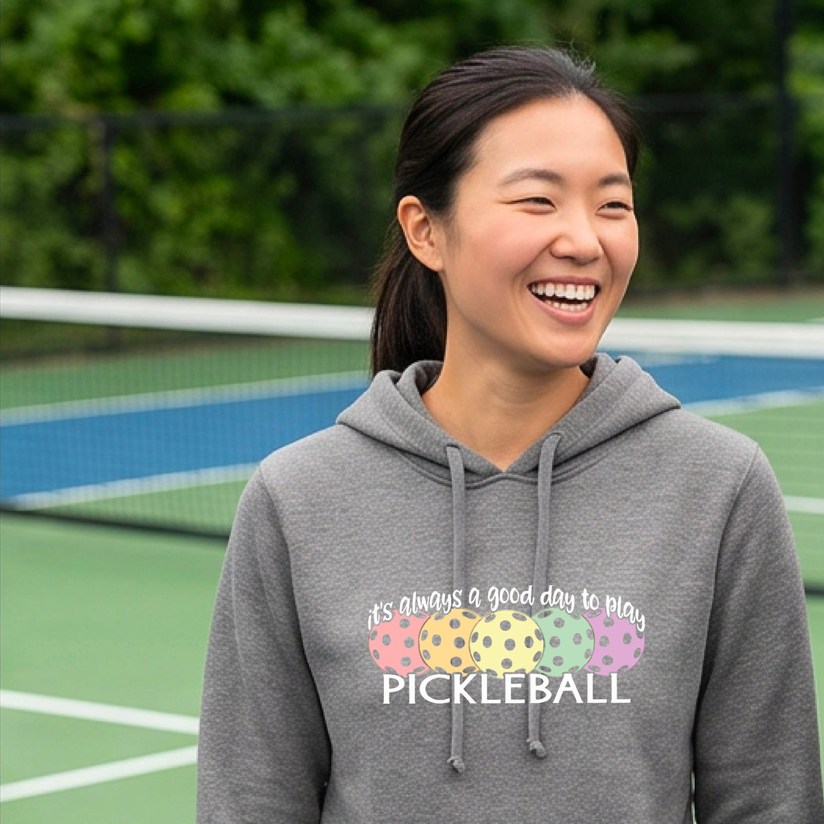 A woman smiling on a pickleball court wearing a gray hoodie with a graphic that reads 'it's always a good day to play PICKLEBALL' above four colorful pickleballs.