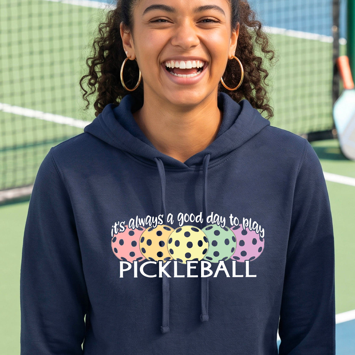 A woman smiling on a pickleball court wearing a navy blue hoodie with a graphic that reads 'it's always a good day to play PICKLEBALL' above four colorful pickleballs.