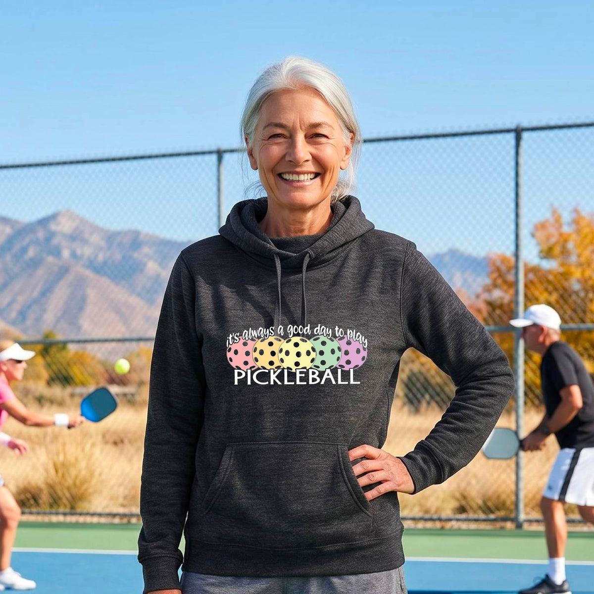 A smiling woman stands on a pickleball court wearing a heather black hoodie with a colorful graphic that reads 'it's always a good day to play PICKLEBALL'. with colorful pickleball graphics. 