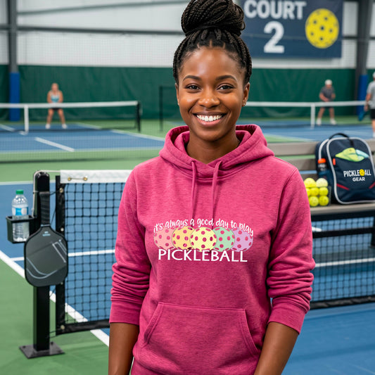 A smiling woman stands on a pickleball court wearing a pink hoodie with a colorful graphic that reads 'it's always a good day to play PICKLEBALL'. with colorful pickleball graphics. 