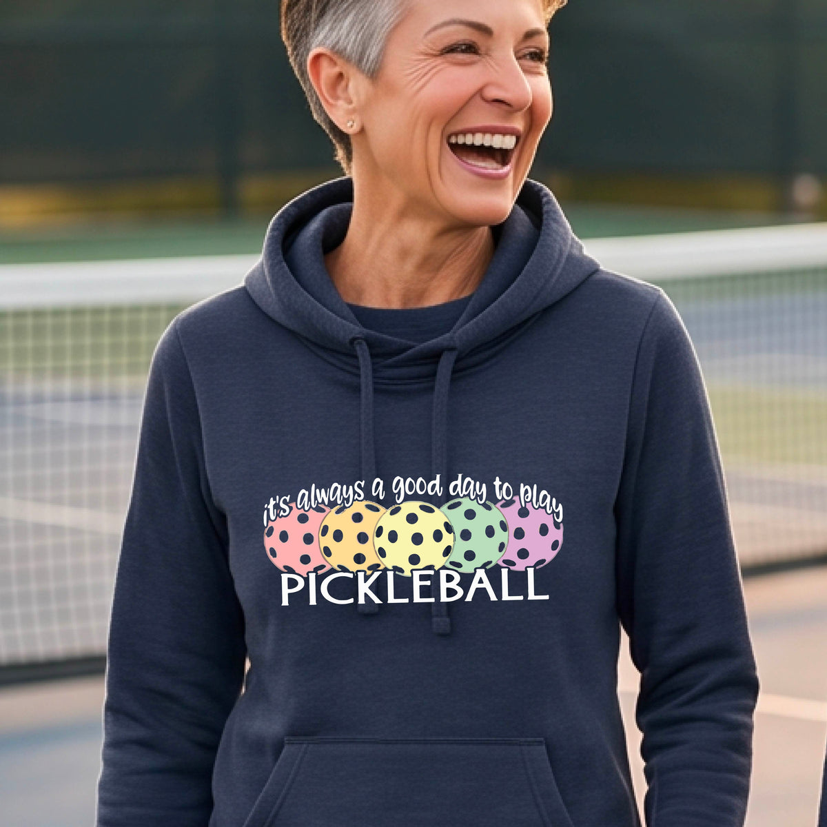 A smiling woman stands on a pickleball court wearing a navy blue hoodie with a colorful graphic that reads 'it's always a good day to play PICKLEBALL'. with colorful pickleball graphics. 