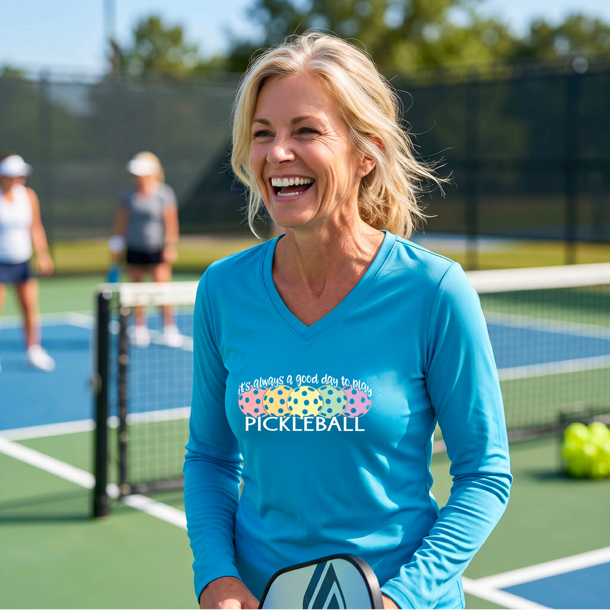 A woman laughing on a pickleball court wearing a light blue long-sleeve V-neck shirt with a colorful pickleball graphic and the text 'it's always a good day to play PICKLEBALL'.