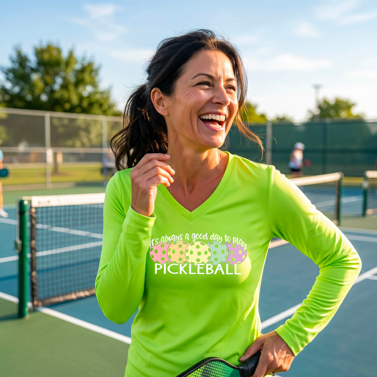 A woman laughing on a pickleball court wearing a bright lime green long-sleeve V-neck shirt with a colorful pickleball graphic and the text 'it's always a good day to play PICKLEBALL'.