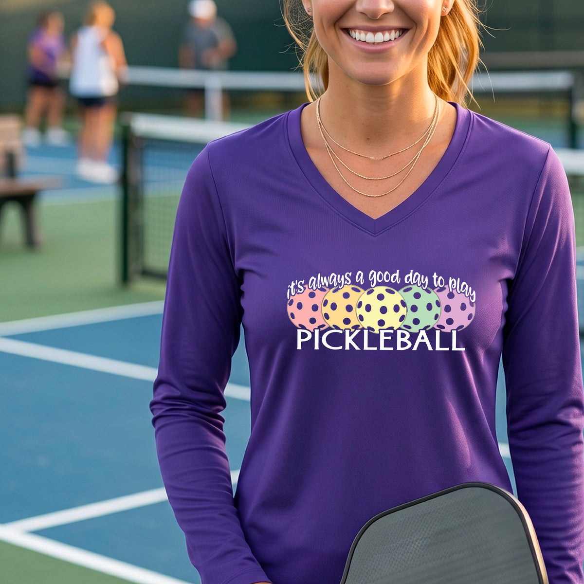 A woman laughing on a pickleball court wearing a purple long-sleeve V-neck shirt with a colorful pickleball graphic and the text 'it's always a good day to play PICKLEBALL'.