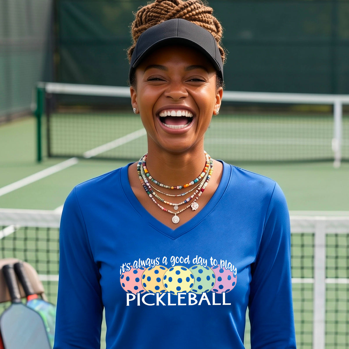 A woman laughing on a pickleball court wearing a blue long-sleeve V-neck shirt with a colorful pickleball graphic and the text 'it's always a good day to play PICKLEBALL'.