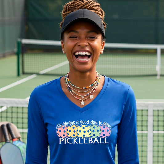 A woman laughing on a pickleball court wearing a blue long-sleeve V-neck shirt with a colorful pickleball graphic and the text 'it's always a good day to play PICKLEBALL'.