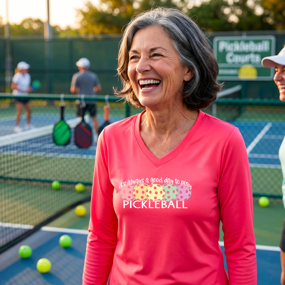 A woman laughing on a pickleball court wearing a bright coral pink long-sleeve V-neck shirt with a colorful pickleball graphic and the text 'it's always a good day to play PICKLEBALL'.