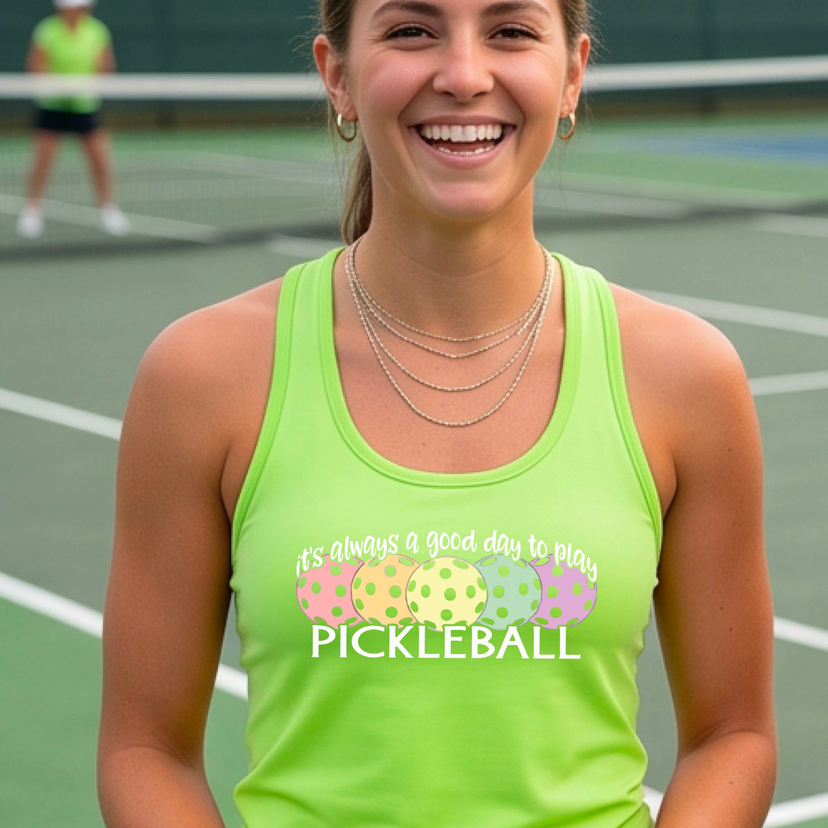 A woman n a pickleball court wearing a bright lime green  racerback tank top with a colorful pickleball graphic and the text 'It's always a good day to play PICKLEBALL'.