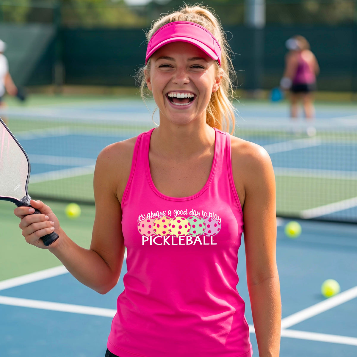 A woman n a pickleball court wearing a hot pink  racerback tank top with a colorful pickleball graphic and the text 'It's always a good day to play PICKLEBALL'.