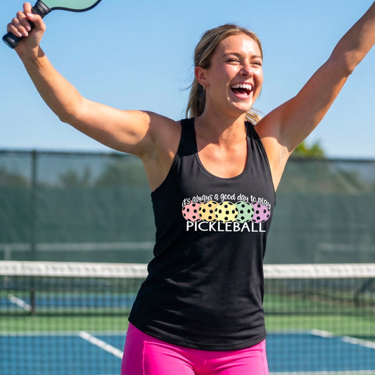 A woman celebrating on a pickleball court wearing a black racerback tank top with a colorful pickleball graphic and the text 'It's always a good day to play PICKLEBALL'.