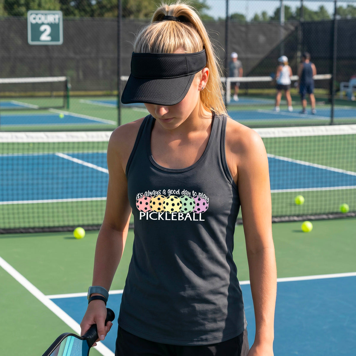 A woman n a pickleball court wearing a hot gray racerback tank top with a colorful pickleball graphic and the text 'It's always a good day to play PICKLEBALL'.