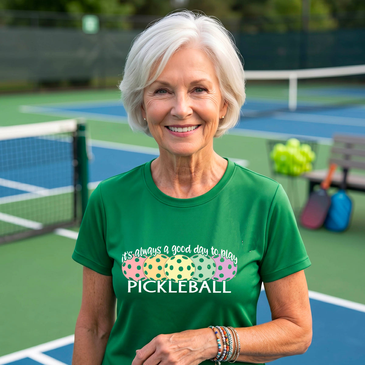 A woman wearing a green short-sleeve t-shirt with a colorful graphic that reads 'it's always a good day to play PICKLEBALL' above five polka-dotted pickleballs. The background is a blurred pickleall court.