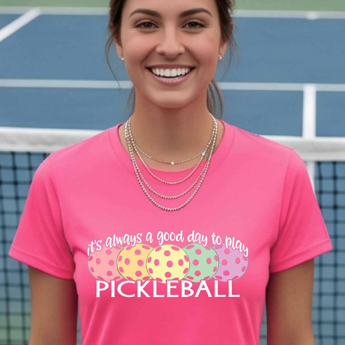 A woman wearing a right coral pink short-sleeve t-shirt with a colorful graphic that reads 'it's always a good day to play PICKLEBALL' above five polka-dotted pickleballs. The background is a blurred pickleall court.