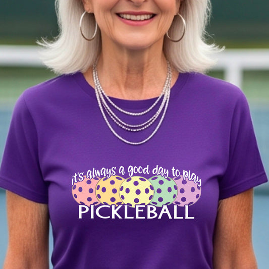 A woman wearing a purple short-sleeve t-shirt with a colorful graphic that reads 'it's always a good day to play PICKLEBALL' above five polka-dotted pickleballs. The background is a blurred pickleall court.