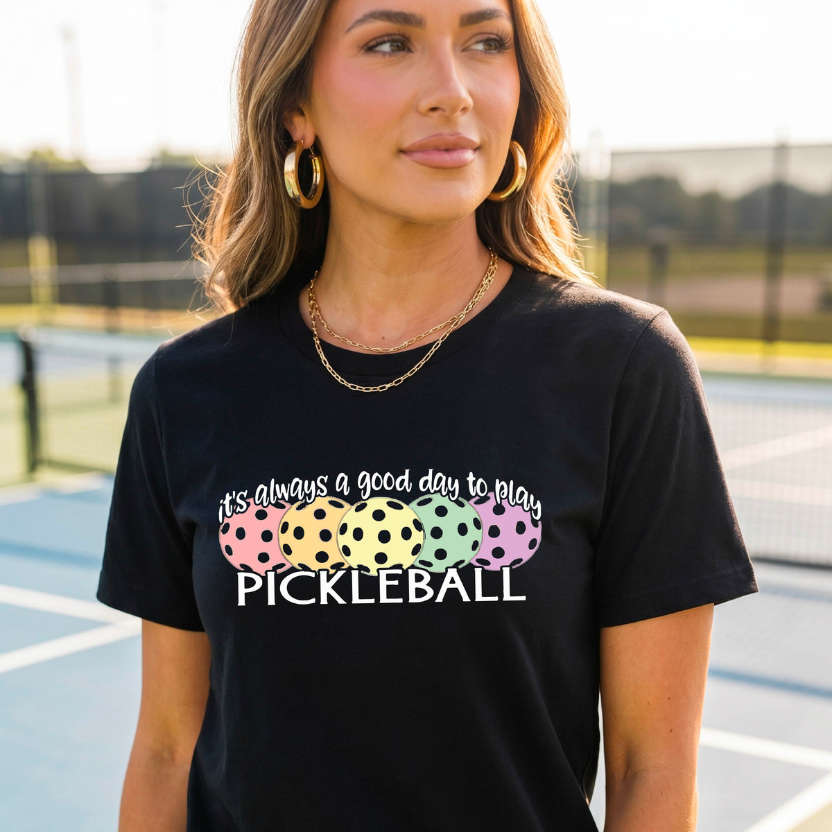 A woman wearing a black short-sleeve t-shirt with a colorful graphic that reads 'it's always a good day to play PICKLEBALL' above five polka-dotted pickleballs. The background is a blurred pickleall court.