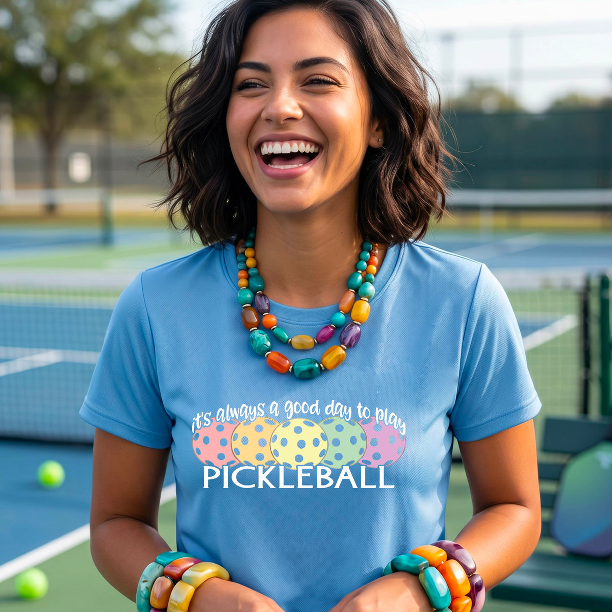 A woman wearing a light blue short-sleeve t-shirt with a colorful graphic that reads 'it's always a good day to play PICKLEBALL' above five polka-dotted pickleballs. The background is a blurred pickleall court.