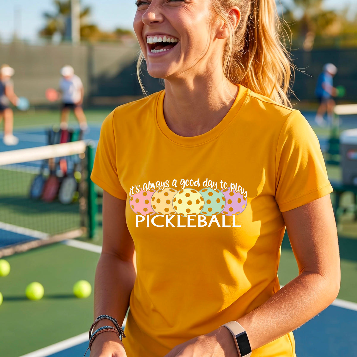 A woman wearing a mustard yellow gold short-sleeve t-shirt with a colorful graphic that reads 'it's always a good day to play PICKLEBALL' above five polka-dotted pickleballs. The background is a blurred pickleall court.