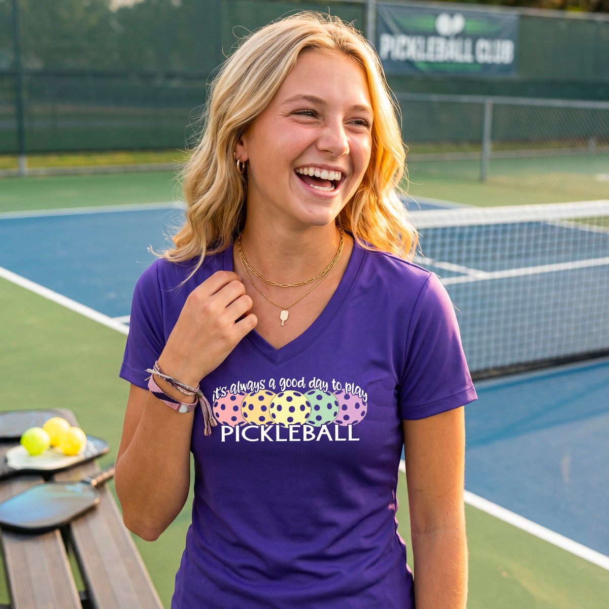 A woman wearing a purple V-neck t-shirt with a colorful graphic that reads 'it's always a good day to play PICKLEBALL' and coloful pickleballs.