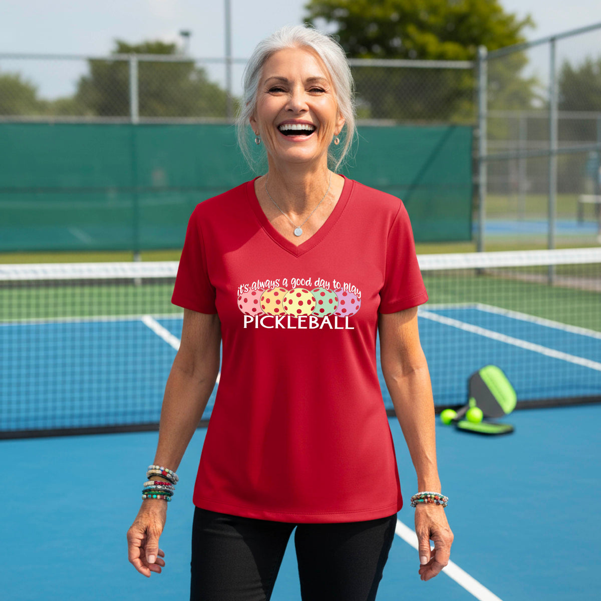 A woman wearing a red V-neck t-shirt with a colorful graphic that reads 'it's always a good day to play PICKLEBALL' and coloful pickleballs.