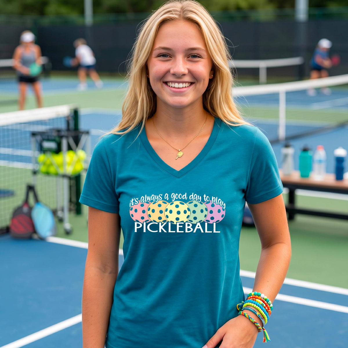 A woman wearing a teal V-neck t-shirt with a colorful graphic that reads 'it's always a good day to play PICKLEBALL' and coloful pickleballs.