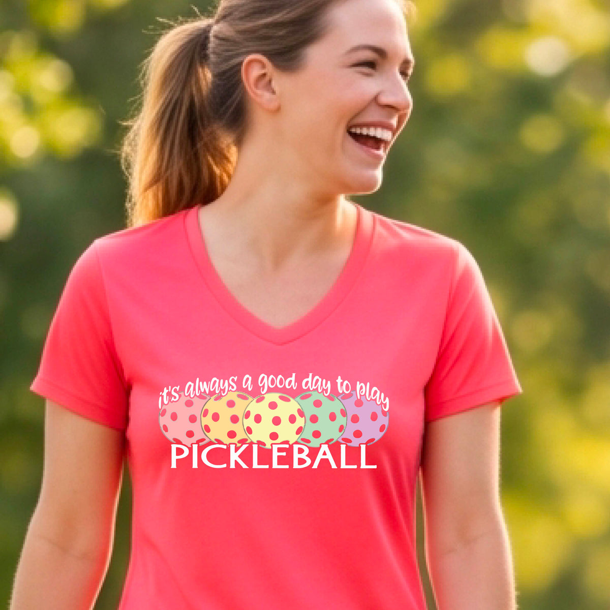 A woman wearing a bright pink V-neck t-shirt with a colorful graphic that reads 'it's always a good day to play PICKLEBALL' and coloful pickleballs.