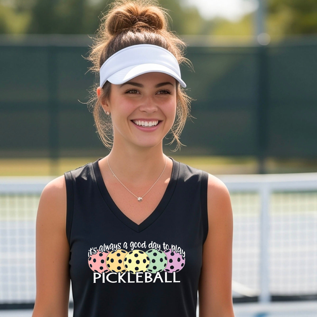 A woman smiling on a pickleball court, wearing a black sleeveless tank top with a colorful pickleball graphic and the text 'it's always a good day to play PICKLEBALL'.