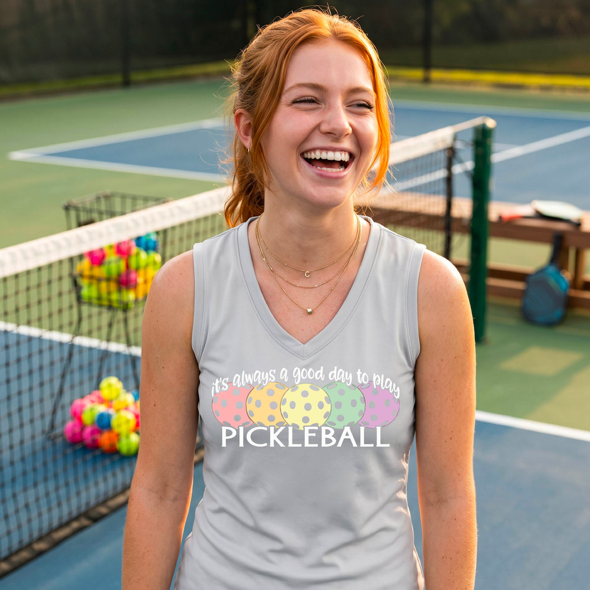 A woman smiling on a pickleball court, wearing a light gray silver sleeveless tank top with a colorful pickleball graphic and the text 'it's always a good day to play PICKLEBALL'.