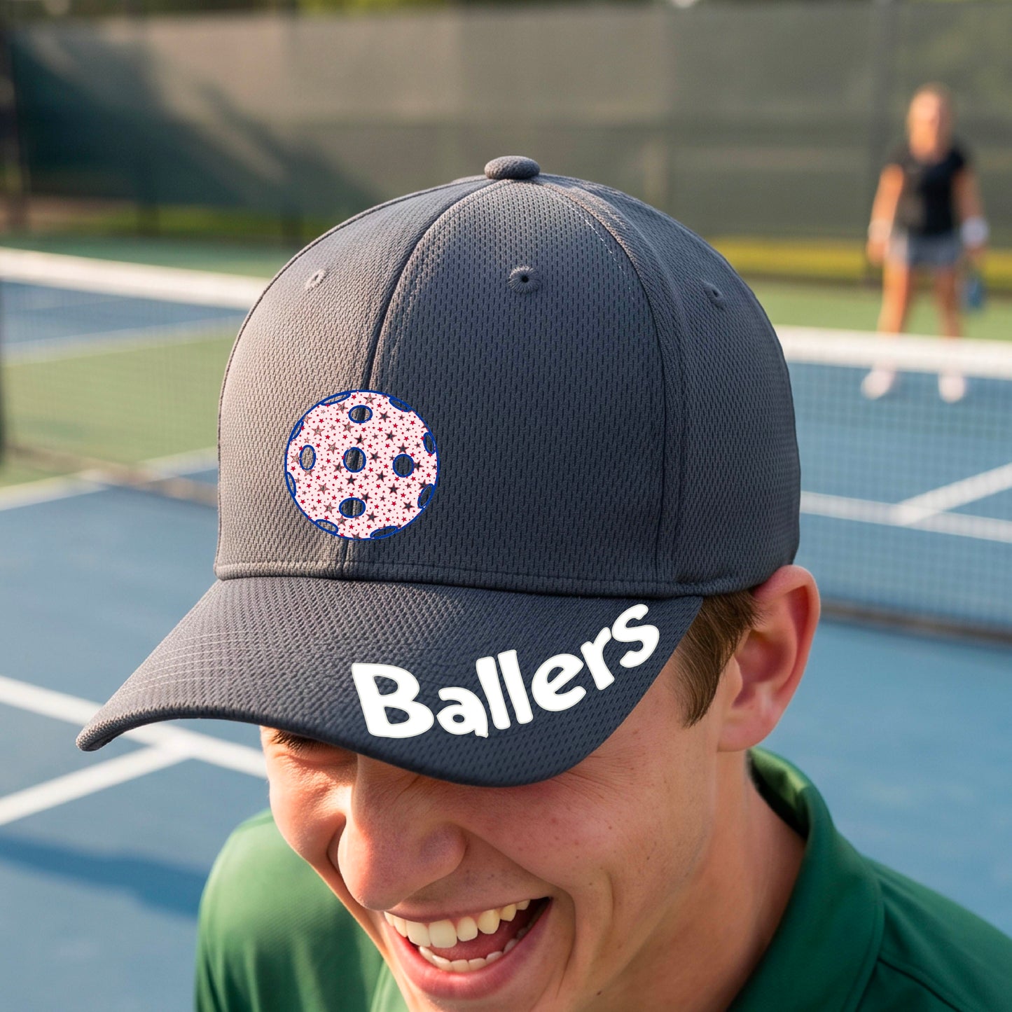 A person on a pickleball court wearing a dark gray baseball cap with a pickleball graphic on the front and the word 'Ballers' in white on the brim.